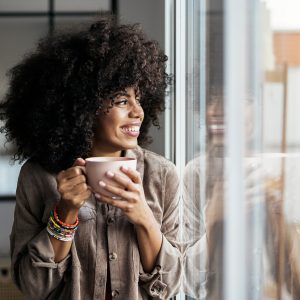 Stock photo of woman having a cup of coffee at home. She is smiling and looking through a window. She looks positive and happy.